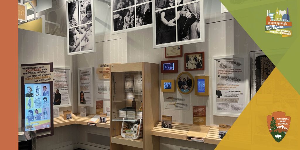 Photograph of a space within the exhibit, featuring old and modern photographs, multicolored text with a glass case displaying personal objects. Digital screens sit awaiting interaction. Around the image stylized shapes and both National Park Service arrowhead logo and city-scape logo labeled One City, Many Cultures in Khmer, English and Spanish.